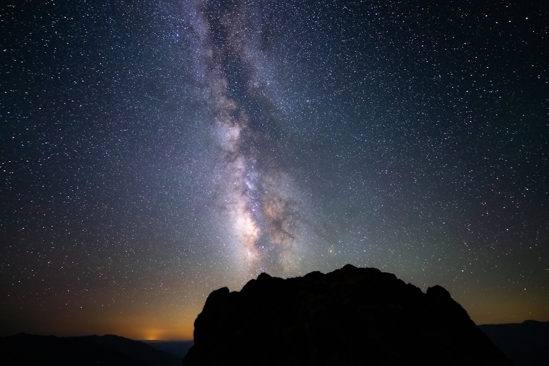 The bright core of the Milky Way rises vertically above the dark silhouette of a rocky peak in Death Valley, filling the sky with countless stars. A dramatic night landscape captured under one of the darkest skies in the American Southwest.